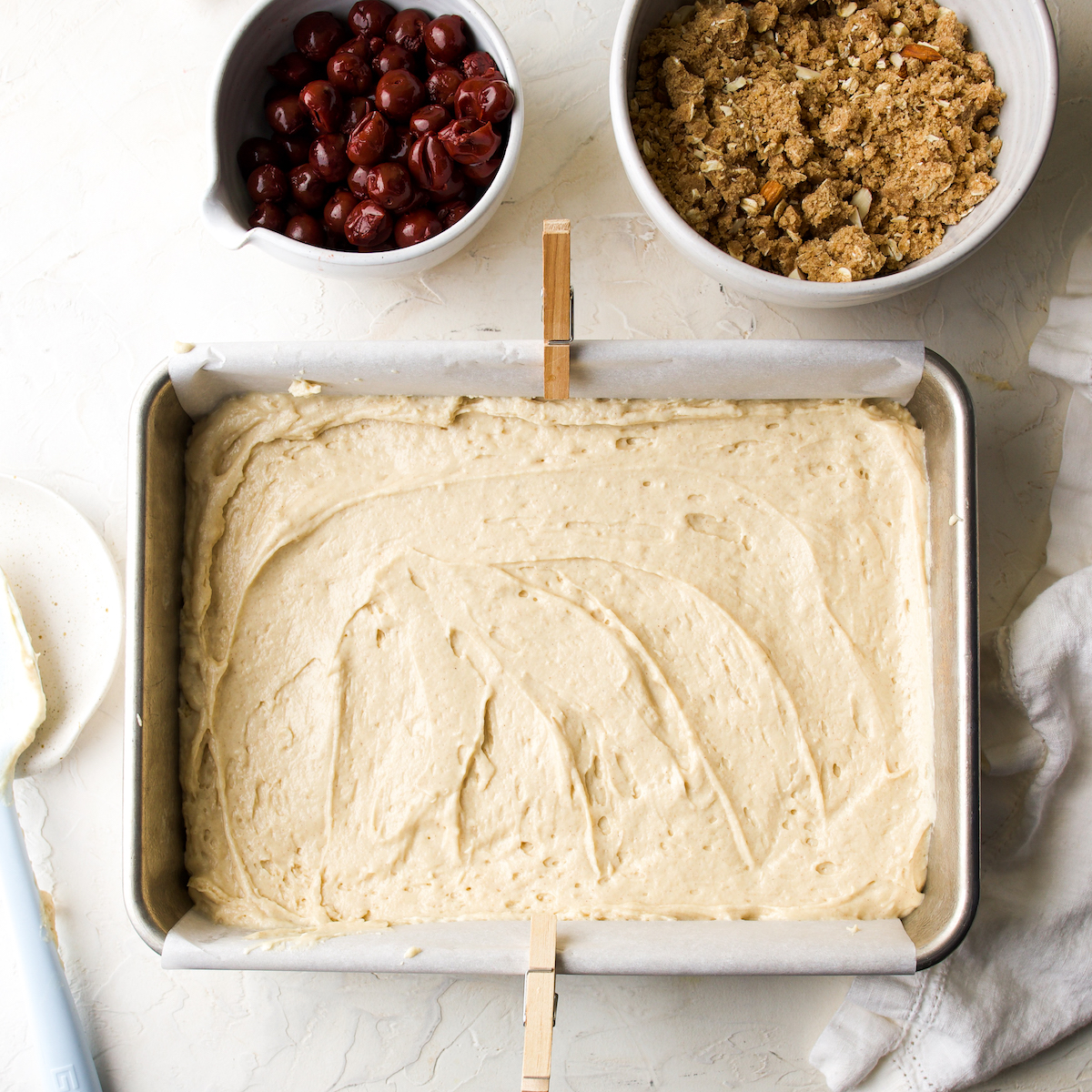 A pan of cake batter next to a bowl of cherries and a bowl of streusel.
