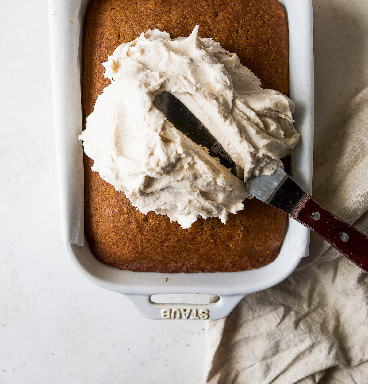 A baking dish with pumpkin cake and buttercream getting smeared on with an offset spatula.