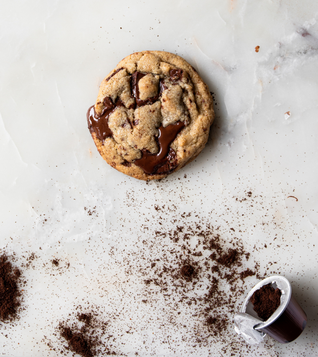 A close up of a chocolate chip cookie with ground espresso on a white marble background.