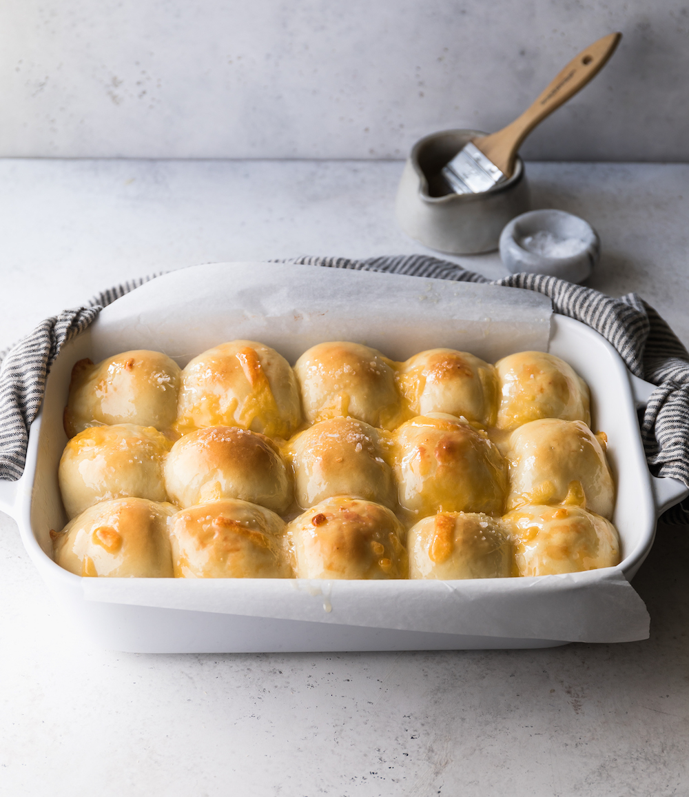 A casserole dish filled with glossy  dinner rolls  on a blueish white background.