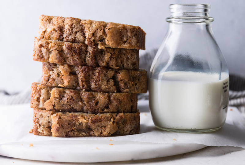 A stack of banana bread next to a glass of milk on a white surface.