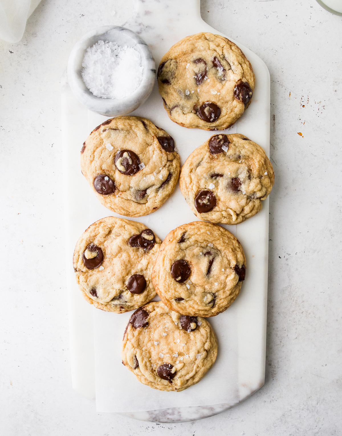A bunch of chocolate chip cookies with a small marble bowl of sea salt on a marble serving board on top of a white surface.