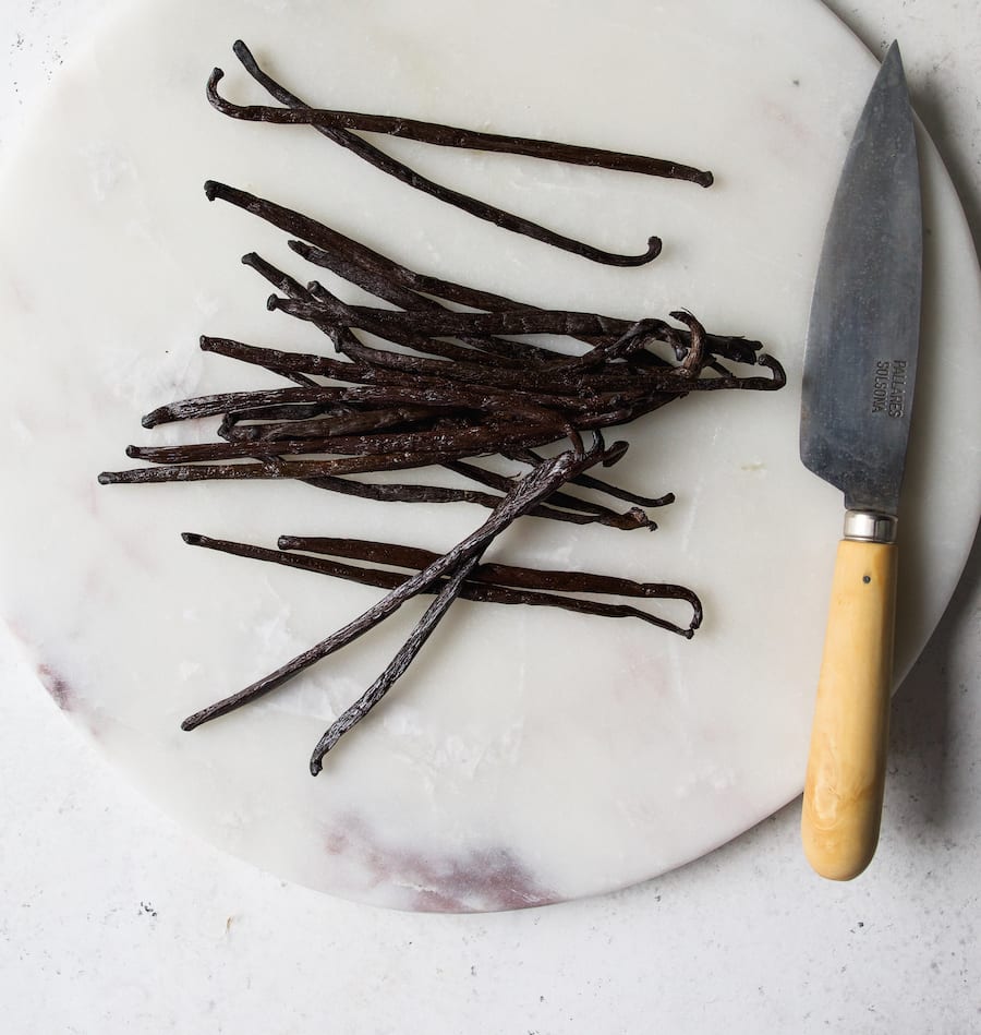A bunch of vanilla bean pods on a marble cutting board with a knife off to the side.