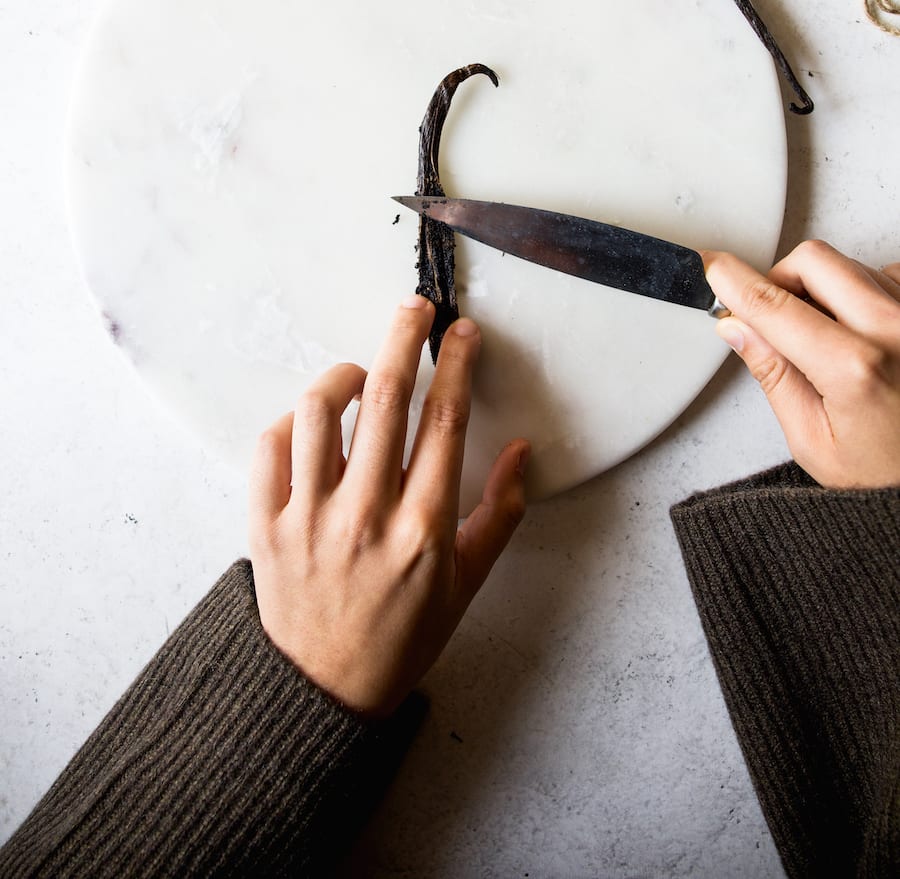 One hand holding a split vanilla bean pod and another hand holding a knife and scraping vanilla bean seeds.