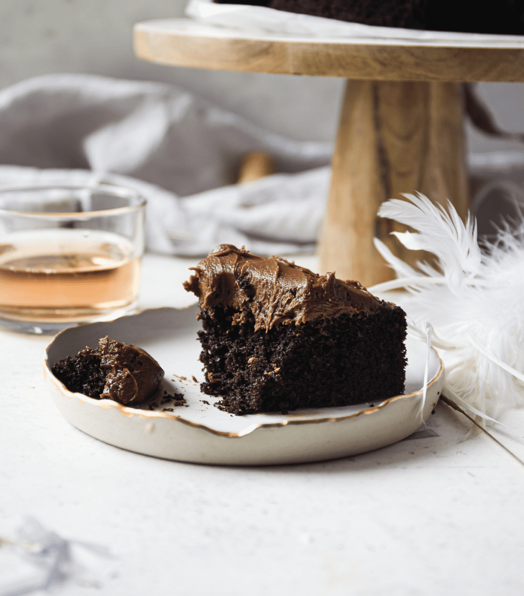 A slice of chocolate cake with chocolate icing on a white plate with a feather beside it