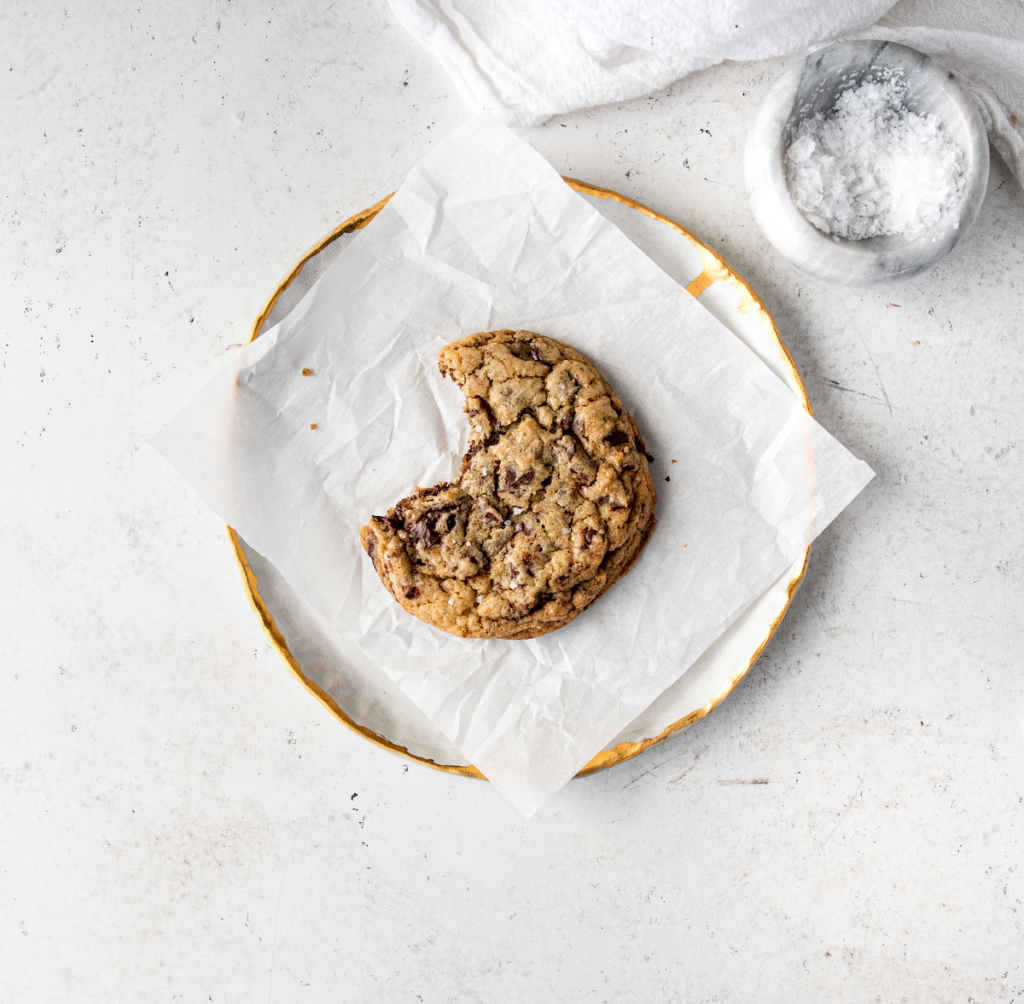 One brown butter chocolate chip cookie on a white plate with a bite out of it.
