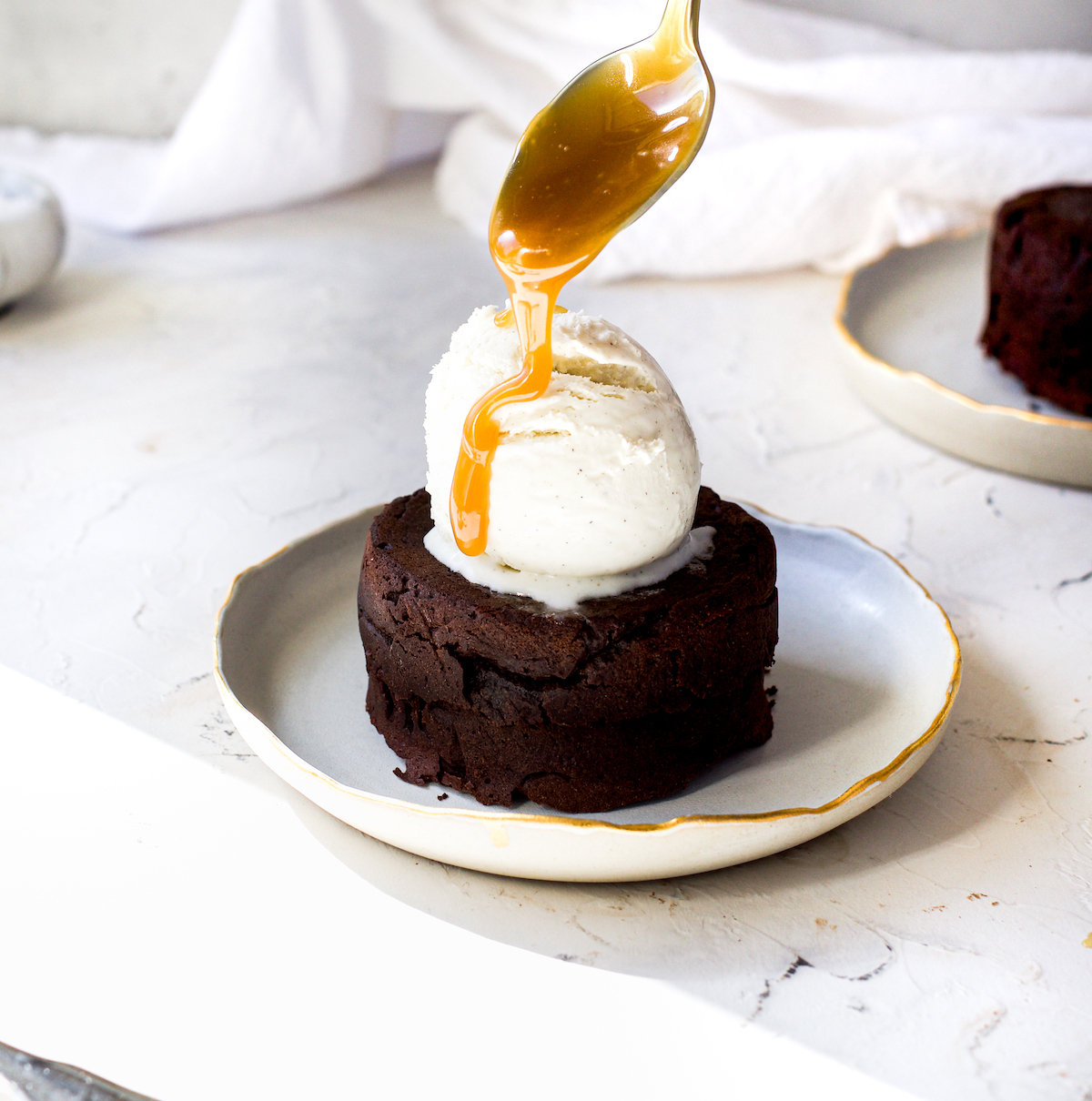 A spoon drizzling caramel onto ice cream on top of a little chocolate cake.