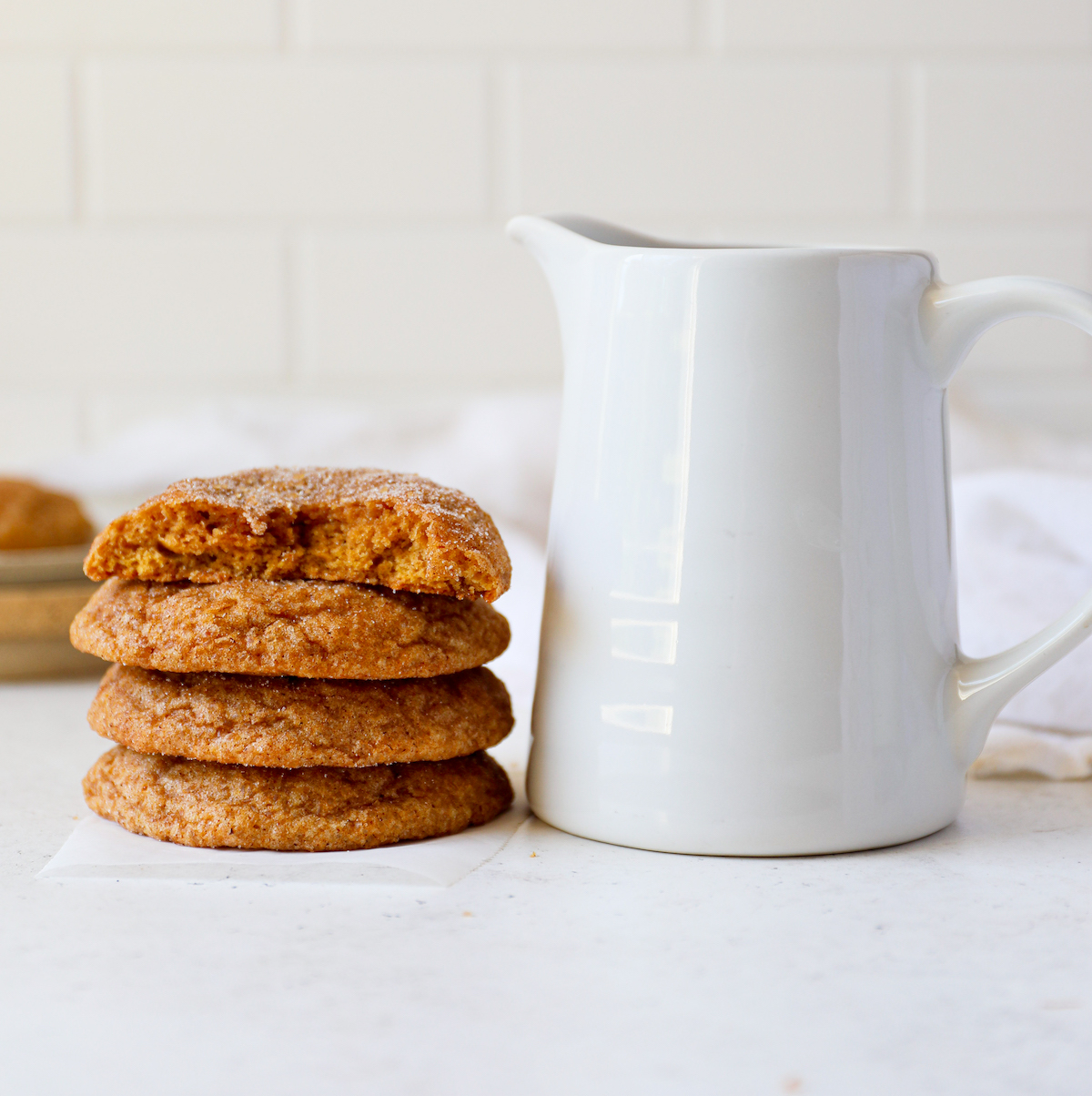 Four stacked pumpkin snickerdoodle cookies next to a milk pitcher on a white surface.