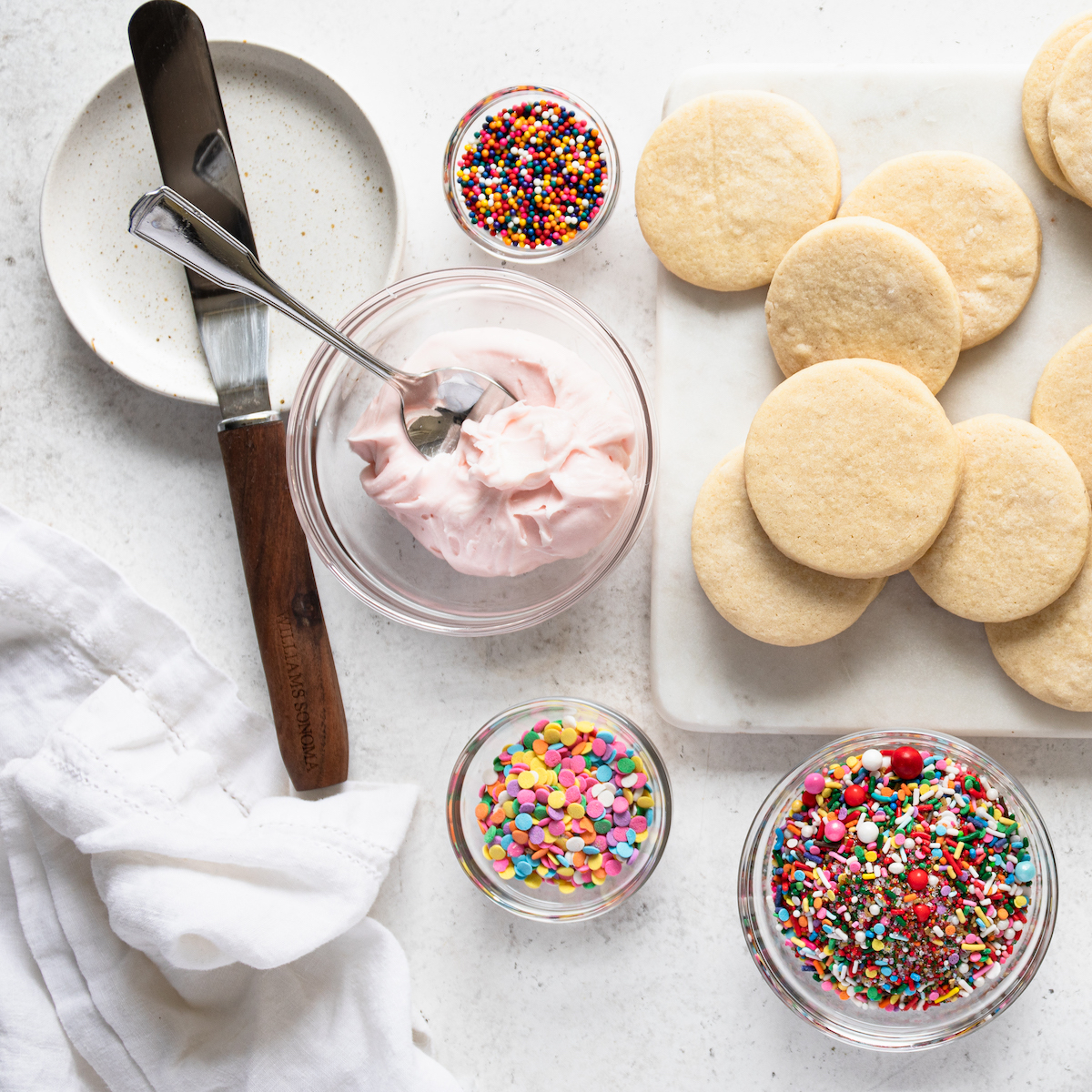 Sugar cookies, sprinkles and vanilla icing on a white surface.