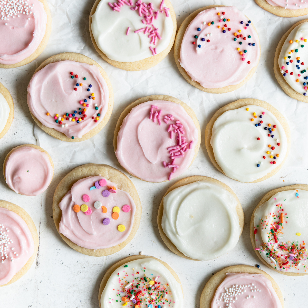A bunch of sugar cookies on a white surface, some with pink frosting and others with white frosting.