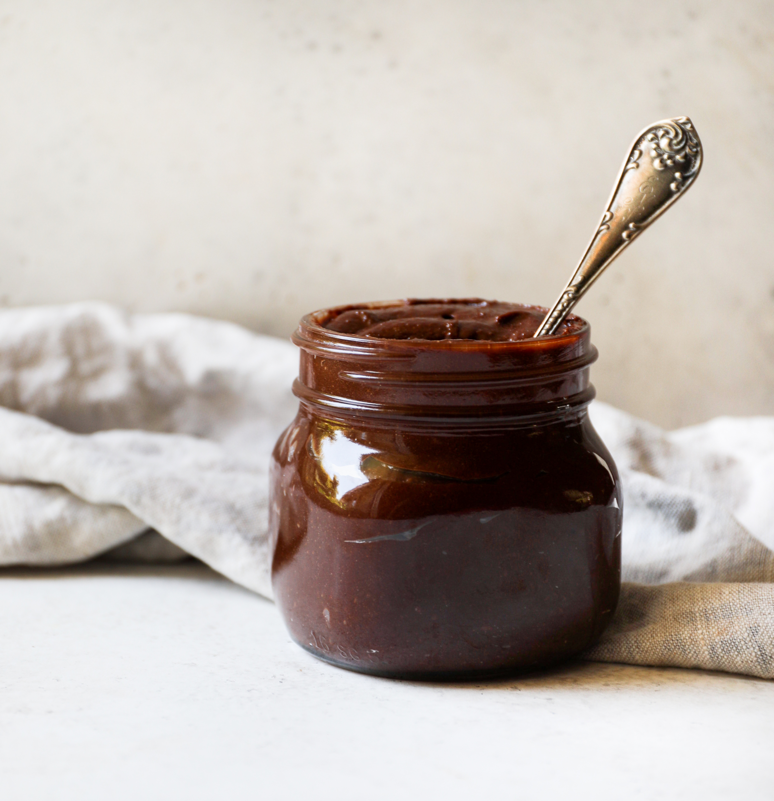 A glass jar set against a white background filled with homemade nutella and a spoon sticking out of it.