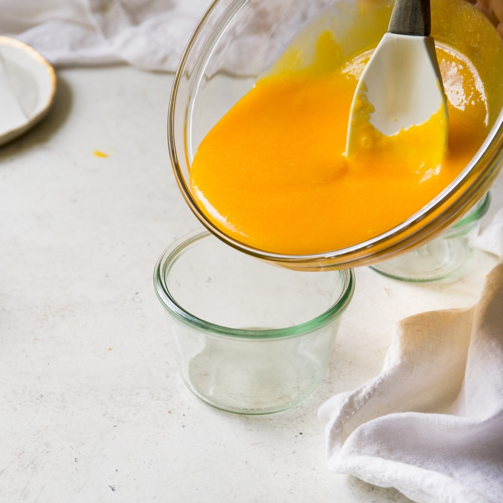 Lemon curd being poured from a glass bowl into a glass jar.