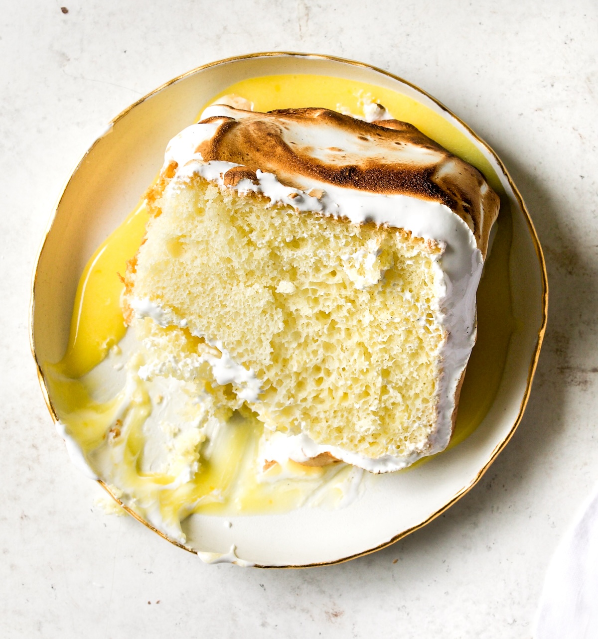 A slice of lemon meringue cake on a white plate on a white background.