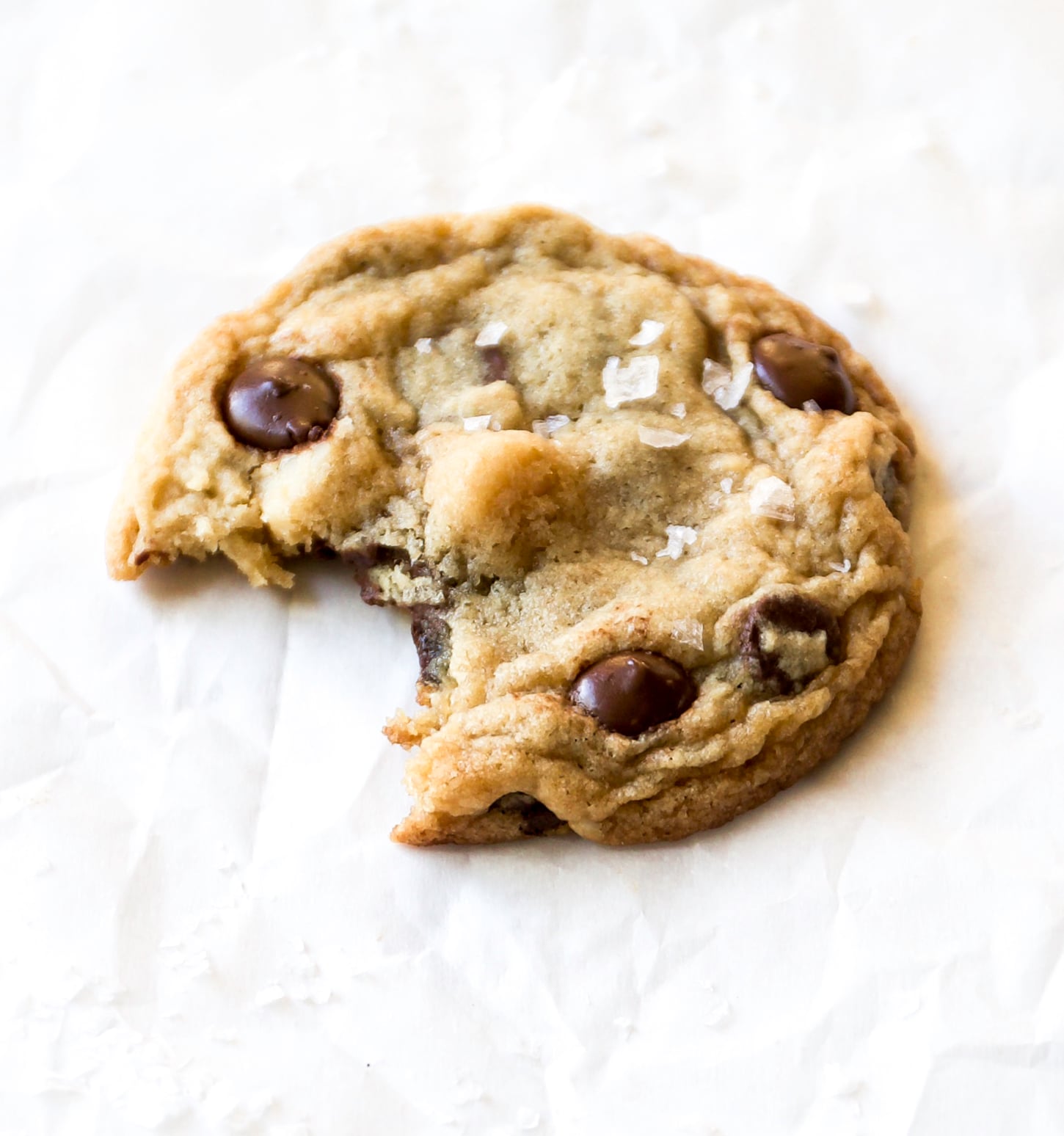 A macadamia nut chocolate chip cookie with a bite taken out of it, sprinkled with flaky sea salt, sits on a white parchment paper background.