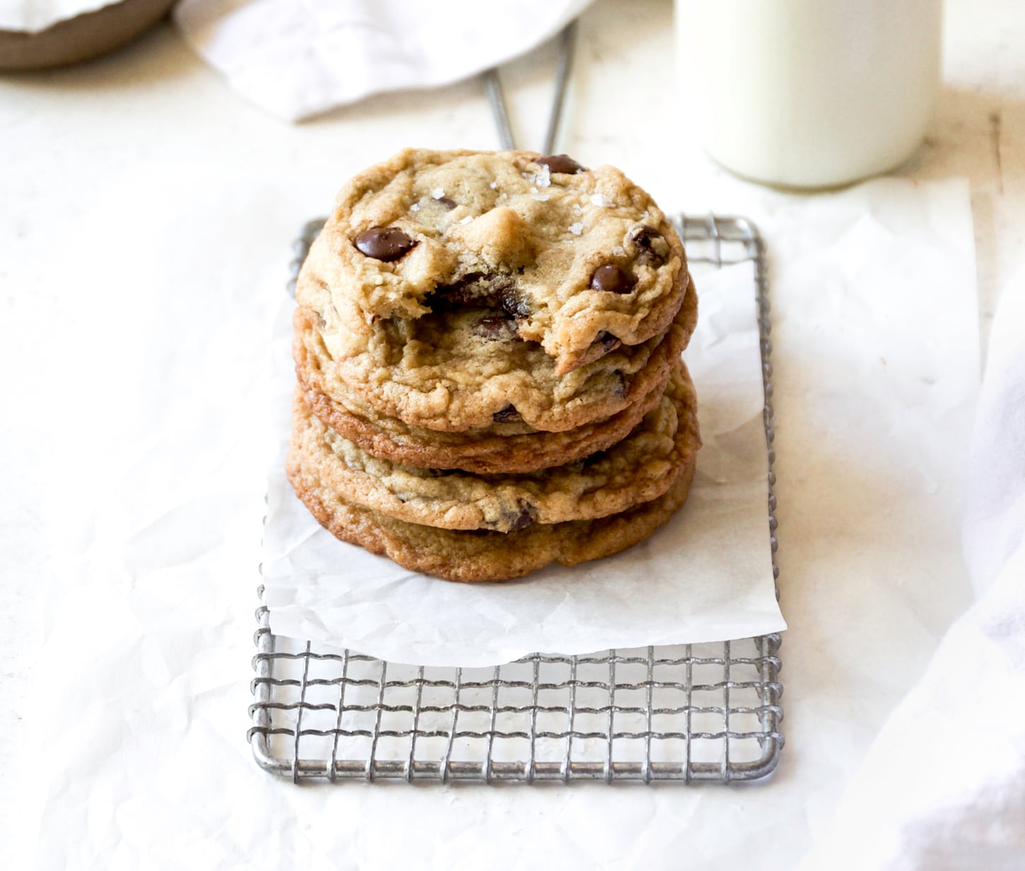 A stack of macadamia nut chocolate chip cookies sits on a small wire cooling rack lined with parchment paper, with a glass of milk in the background.