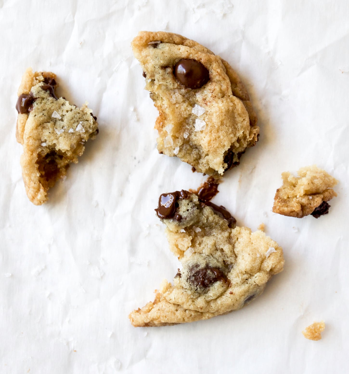 A macadamia nut chocolate chip cookie, broken into several pieces, sits on crumpled white parchment paper, with visible chocolate chips, crunchy macadamia nuts, and a sprinkle of sea salt on top.